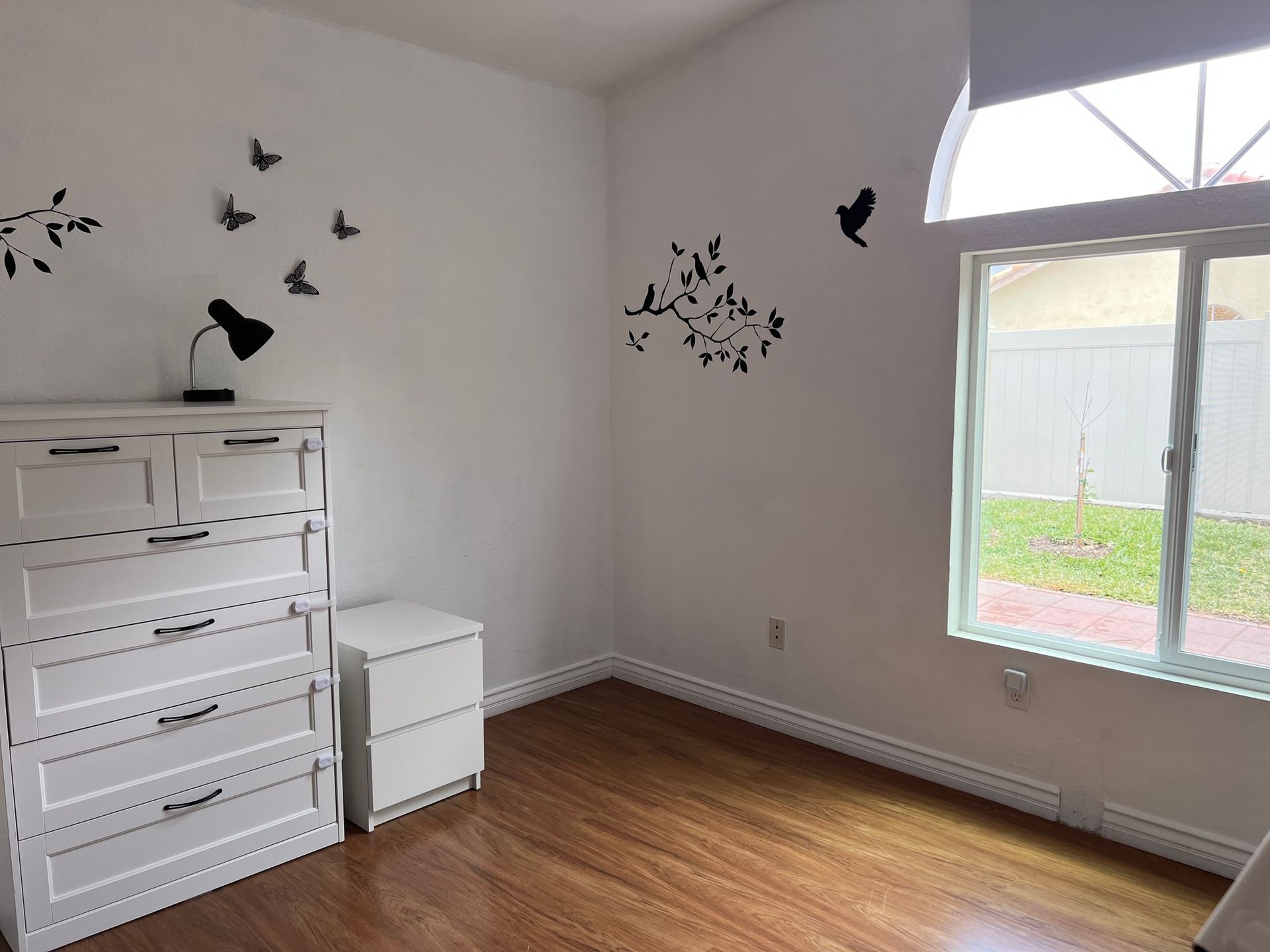 White bedroom with dresser, nightstand, butterflies on wall, window with greenery.