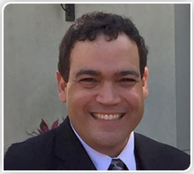 Man in a black suit smiling broadly, dark hair, neutral background, outdoors.