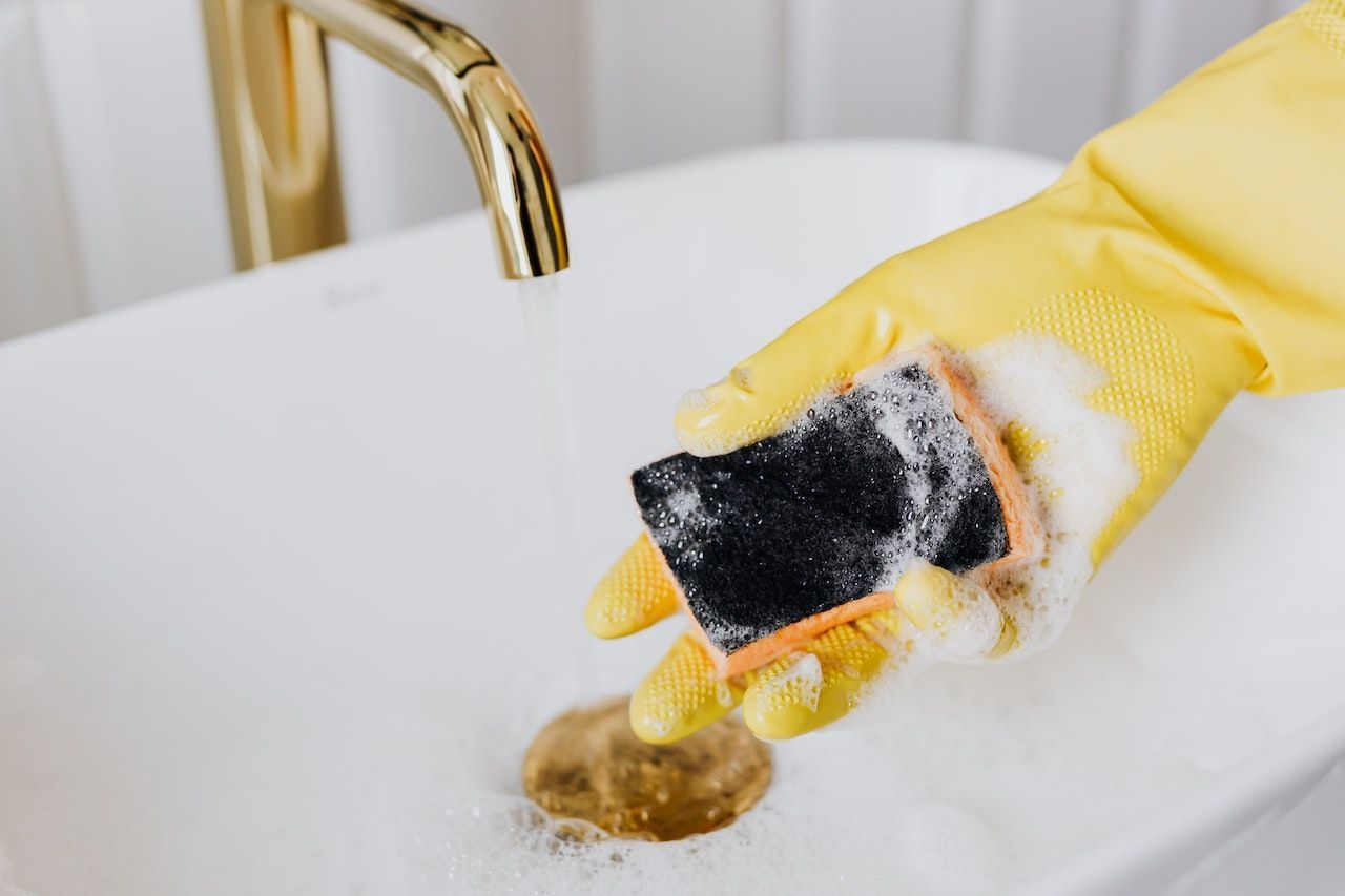 A person wearing yellow gloves is cleaning a sink with a sponge.