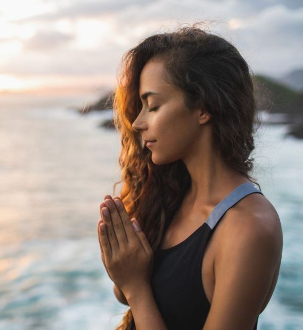 A woman is meditating with her eyes closed in front of the ocean — Essy K Counselling And Wellness Hub In Croudace Bay NSW