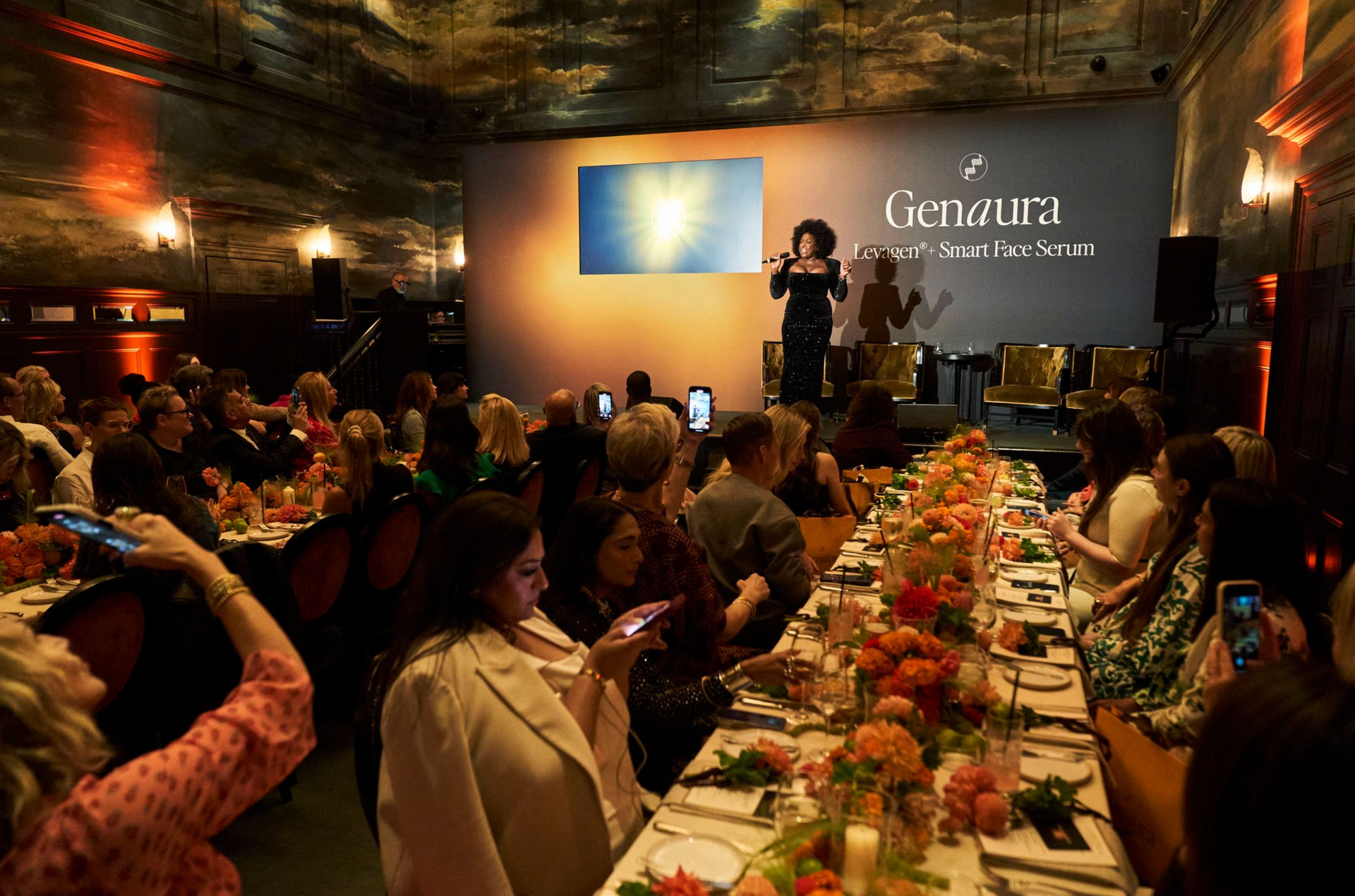 Genaura Launch event, People at a dinner event, speaker on stage with a logo backdrop. Long table with floral arrangements.