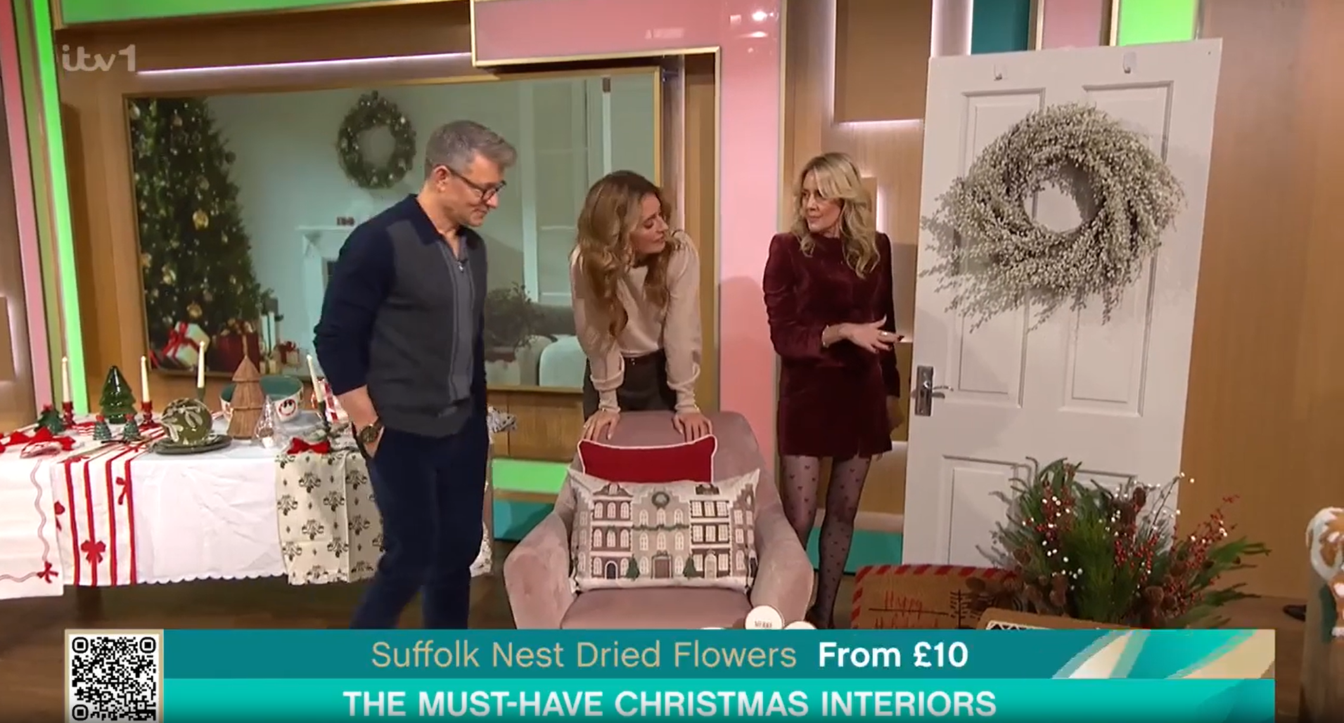 Three presenters stand in a studio decorated for Christmas, showcasing holiday decor, including a wreath on a door.