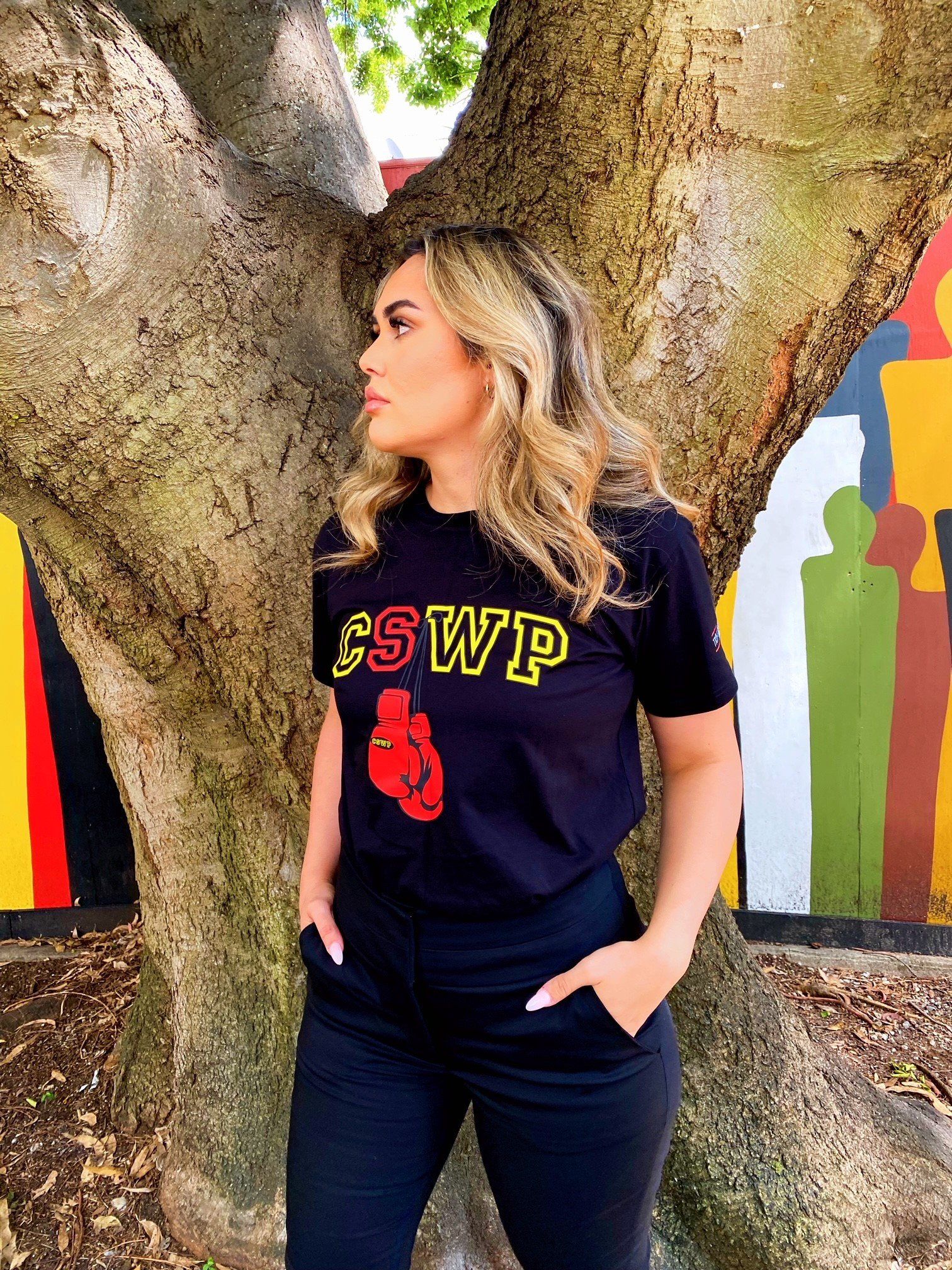 A woman wearing a black cswp t-shirt and black pants is standing in front of a tree.