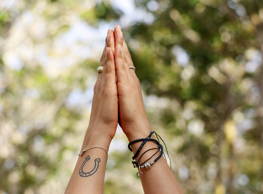 Two hands pressed together in a prayer pose, featuring a horseshoe tattoo on one wrist and bracelets on the other.
