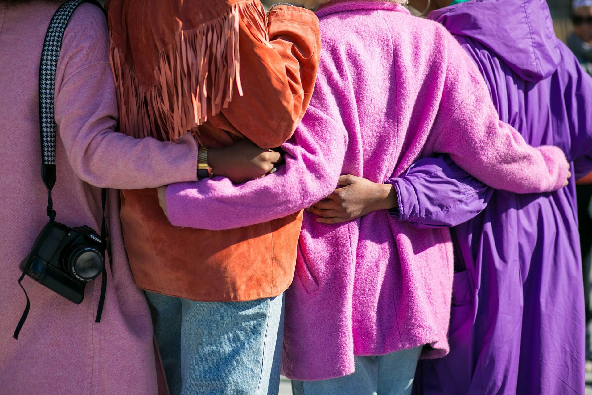 Four people standing in a row, arms around each other, wearing bright pink, orange, and purple jackets.