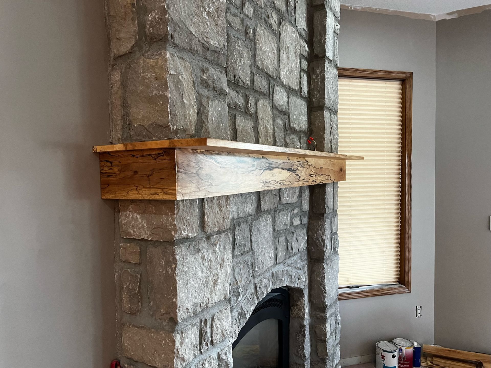 A stone fireplace with a wooden mantle in a living room next to a window.