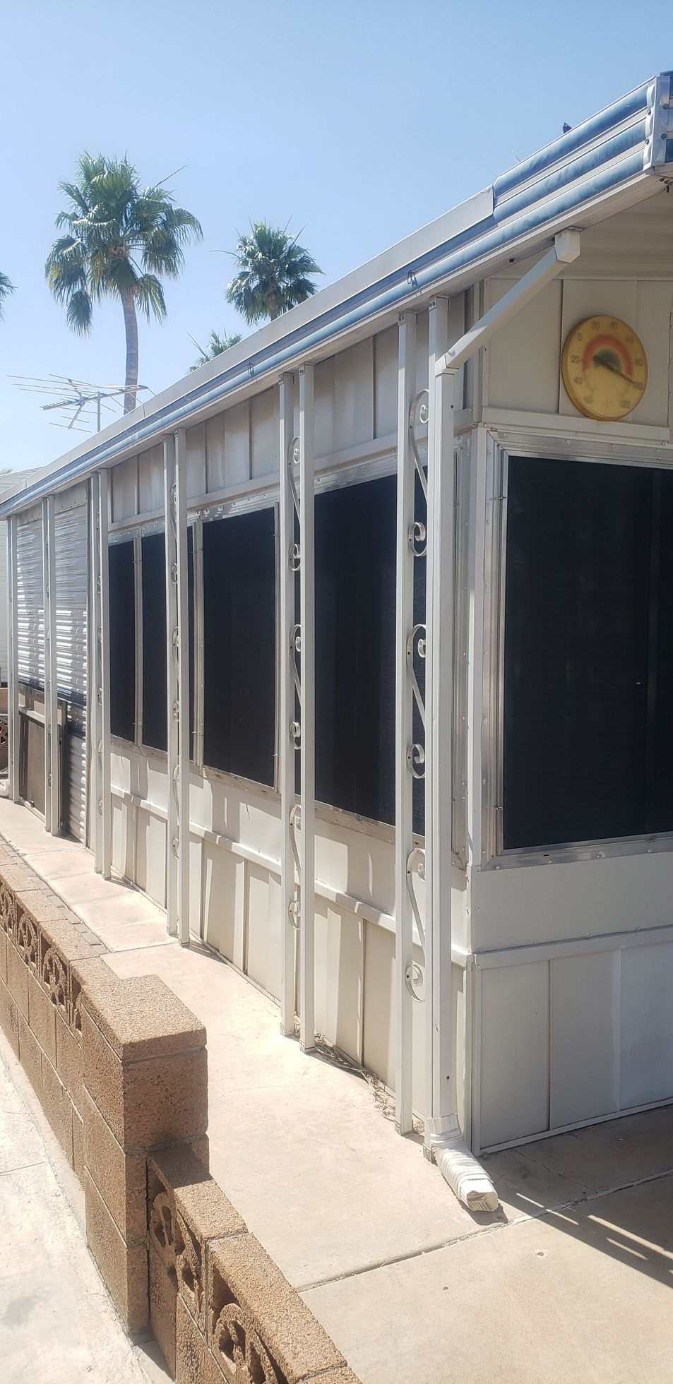 A light-colored building with large dark windows, a concrete walkway, and a brick wall. Palm trees are in the background.