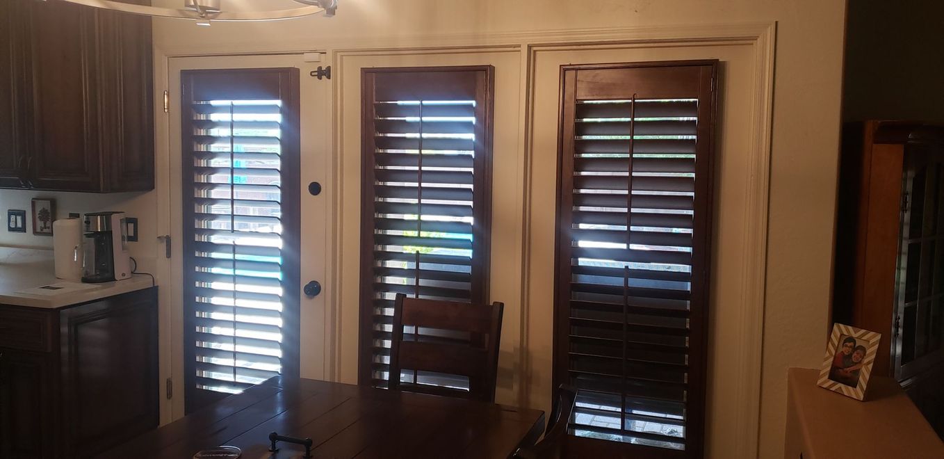 Dark wooden shutters on three windows, with a chair and a table in front. Kitchen setting.