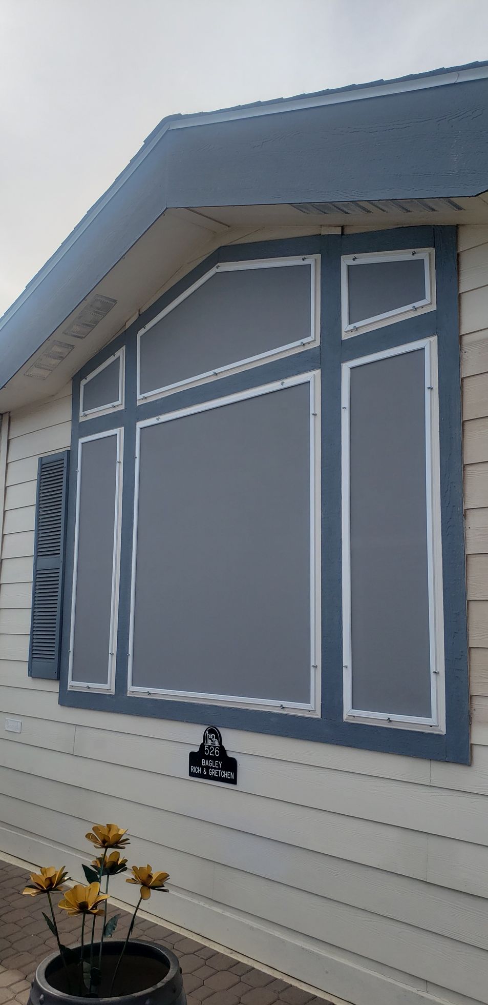 Large window with gray screen on a white house. Blue trim, potted flowers in foreground.