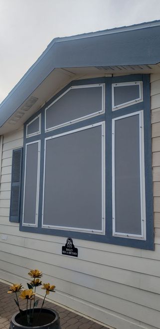 Gray window with white trim on a white house. Blue roof overhang. Flowers in a pot in front.