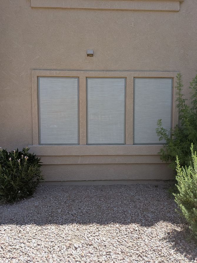 Exterior of a building with three covered windows. Beige stucco wall, gravel ground, and greenery.