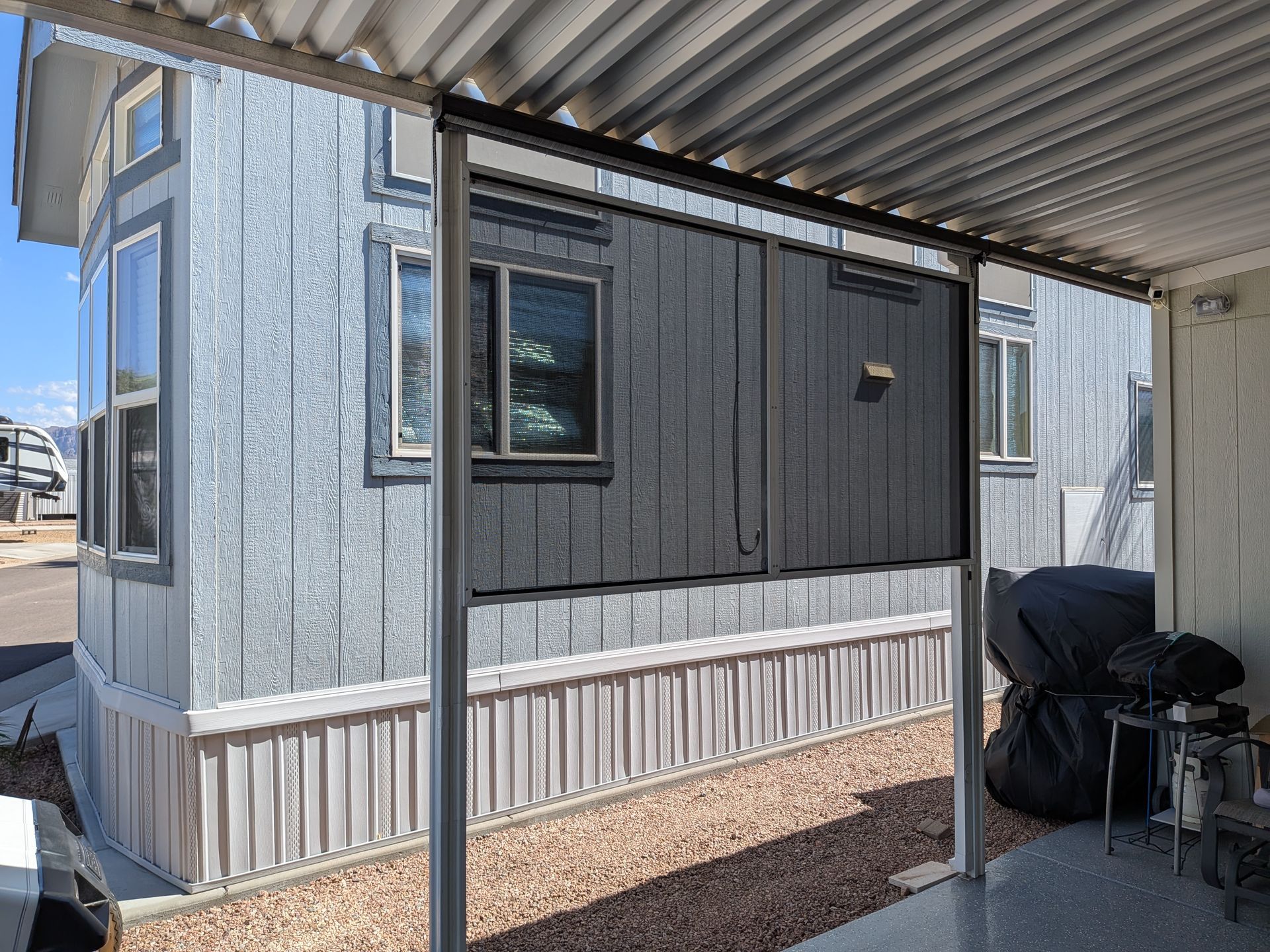 Gray mobile home exterior with porch, gravel ground, and corrugated roof.