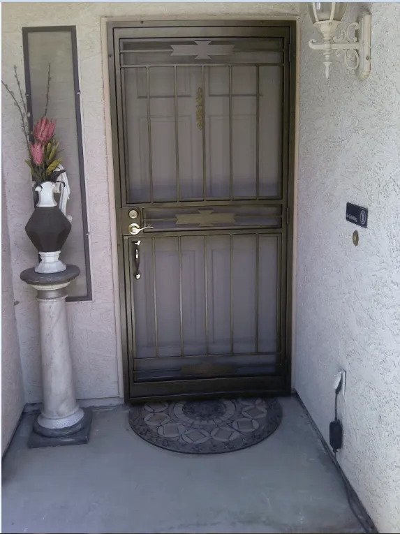 Dark metal security door with doormat, pedestal with vase of flowers, and a light fixture on a stucco wall.