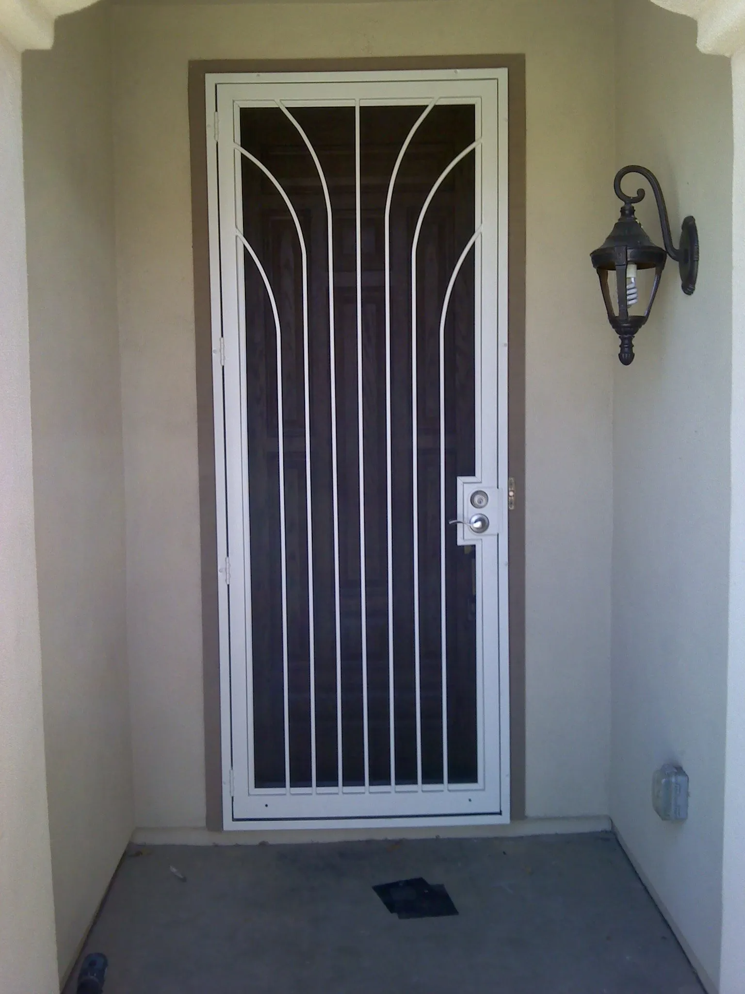 White security door with curved metal bars, on a beige stucco wall, next to a black lantern.