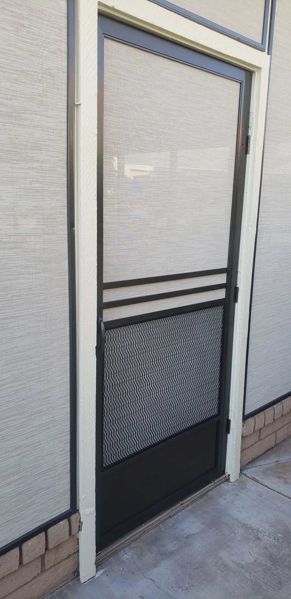 Exterior door with screen; black and grey metal with white trim, set in a textured wall of brick and grey concrete.