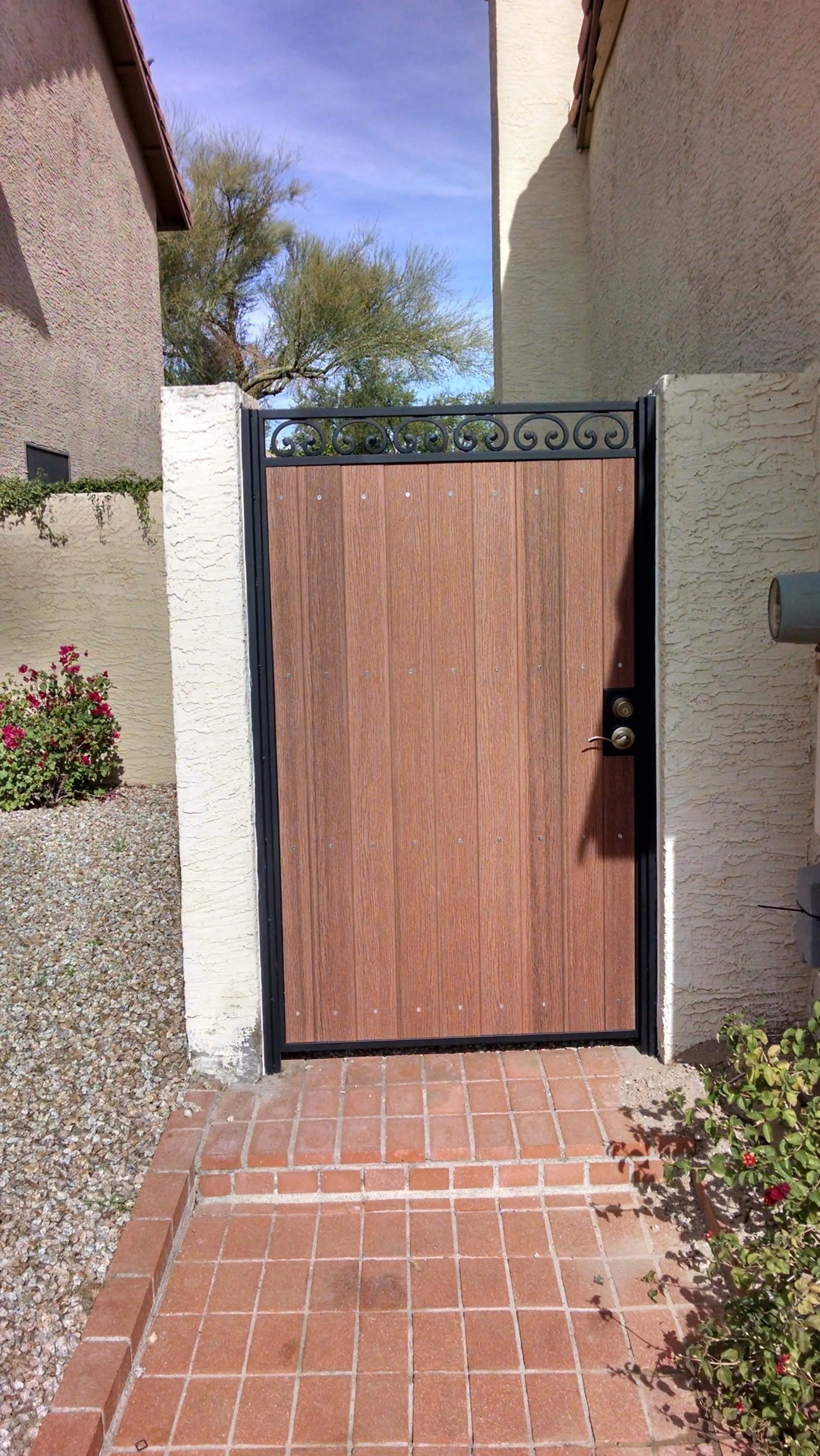 Wooden gate with black metal frame and decorative top, set between stucco walls and brick path.