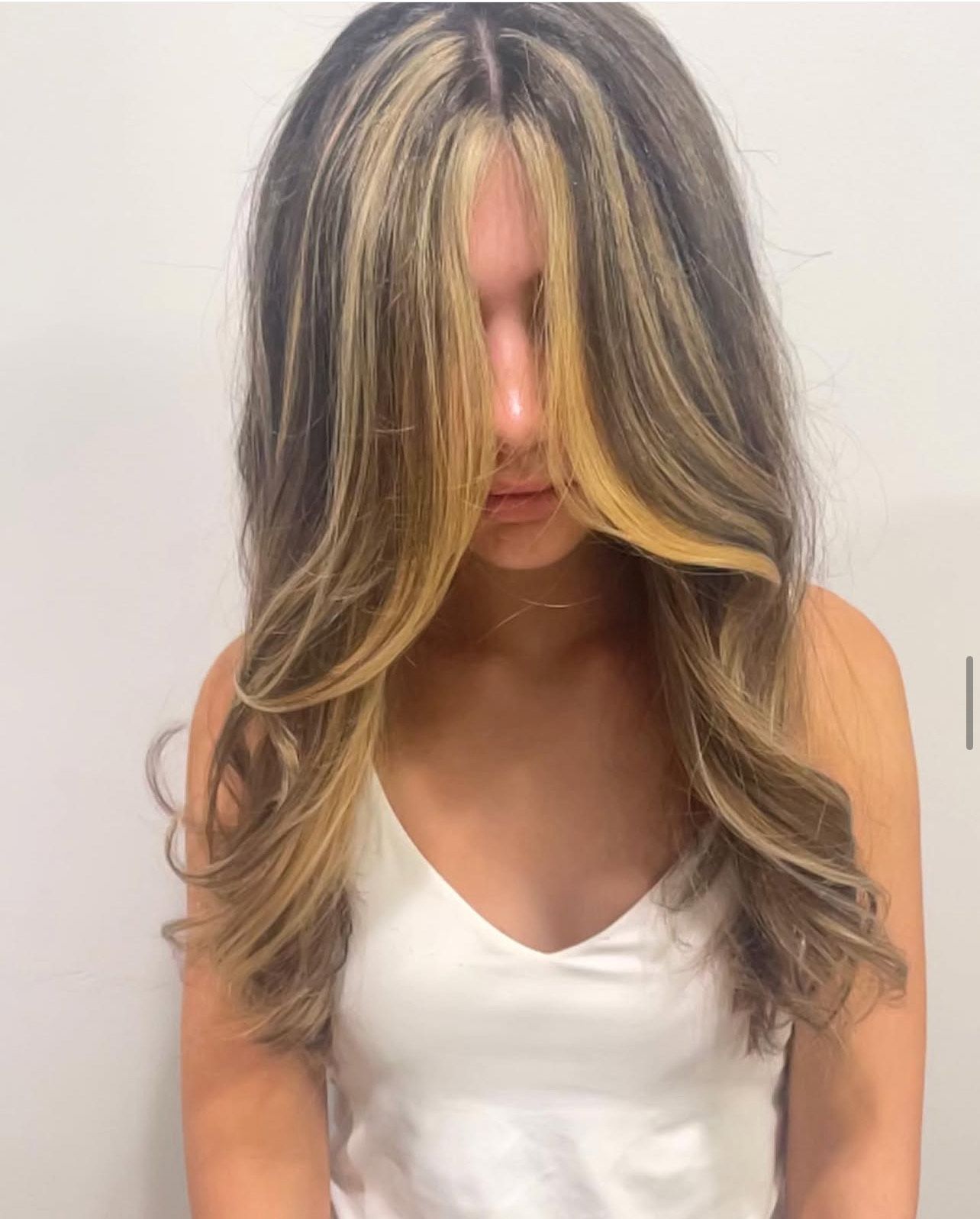 A woman with long brown hair and blonde highlights is wearing a white tank top.