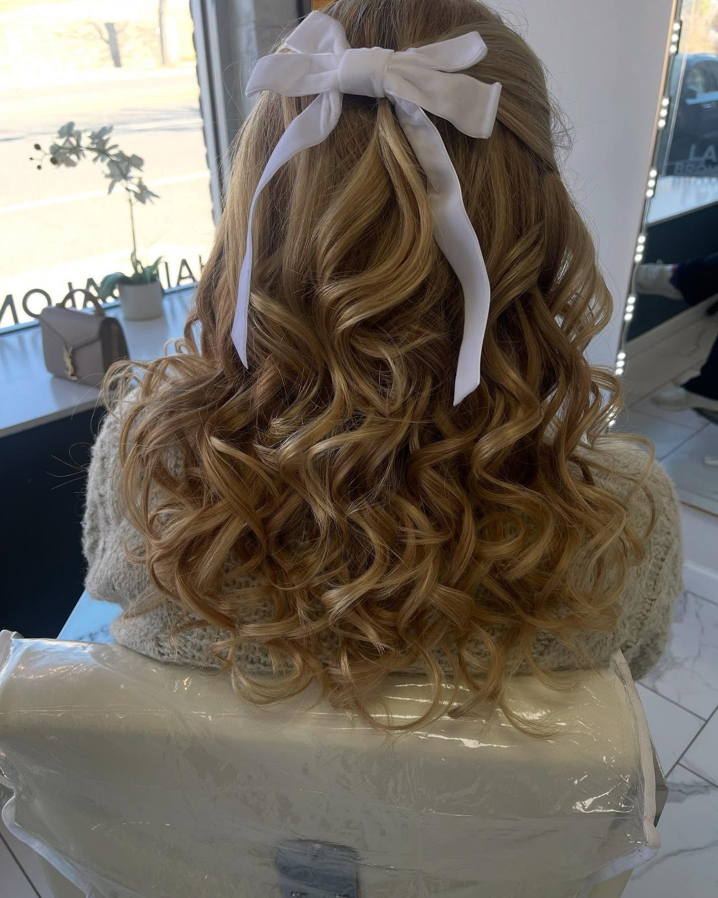 A woman with curly hair is sitting in a chair with a white bow in her hair.