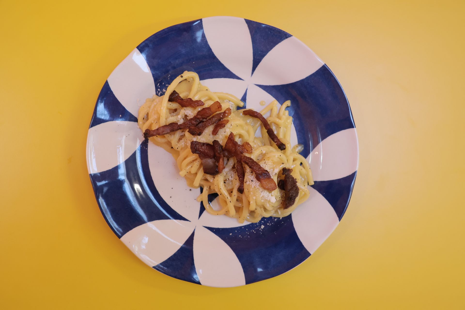 Pasta carbonara with crispy bacon on a blue and white patterned plate on a yellow surface.Domenica 
Home made italian pastries, food & coffee 
