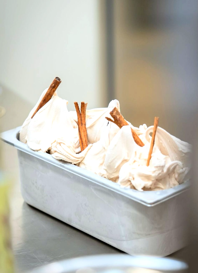 A metal tray of creamy gelato, swirled, topped with cinnamon sticks on a stainless steel surface.