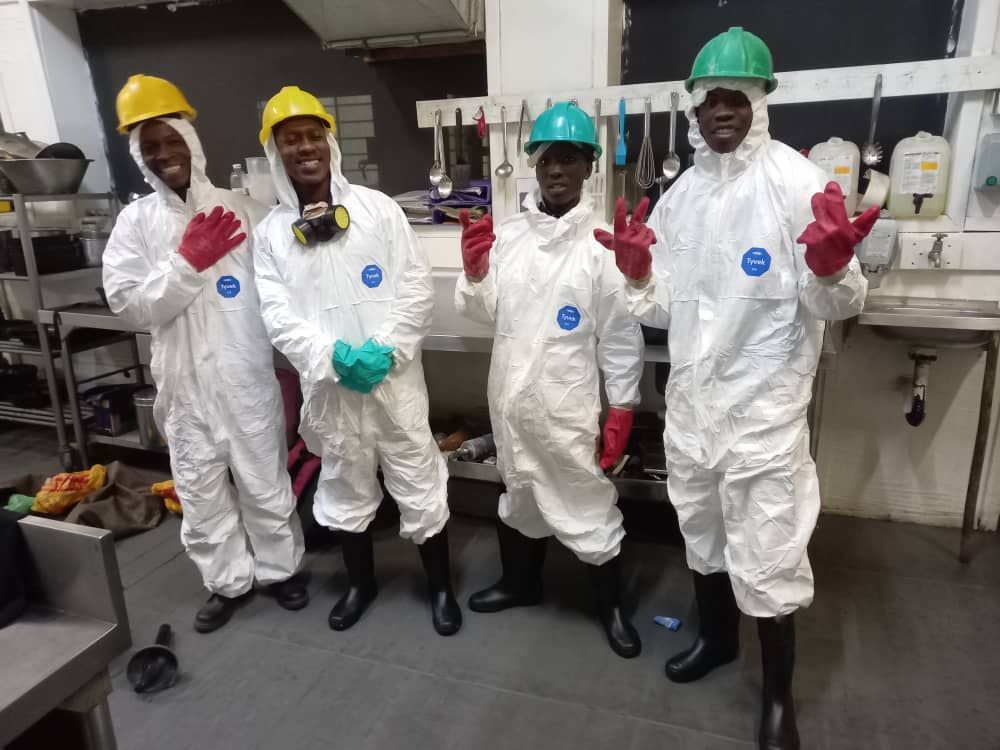 Four people in white hazmat suits, boots, and hard hats, in a kitchen. They gesture with gloved hands.