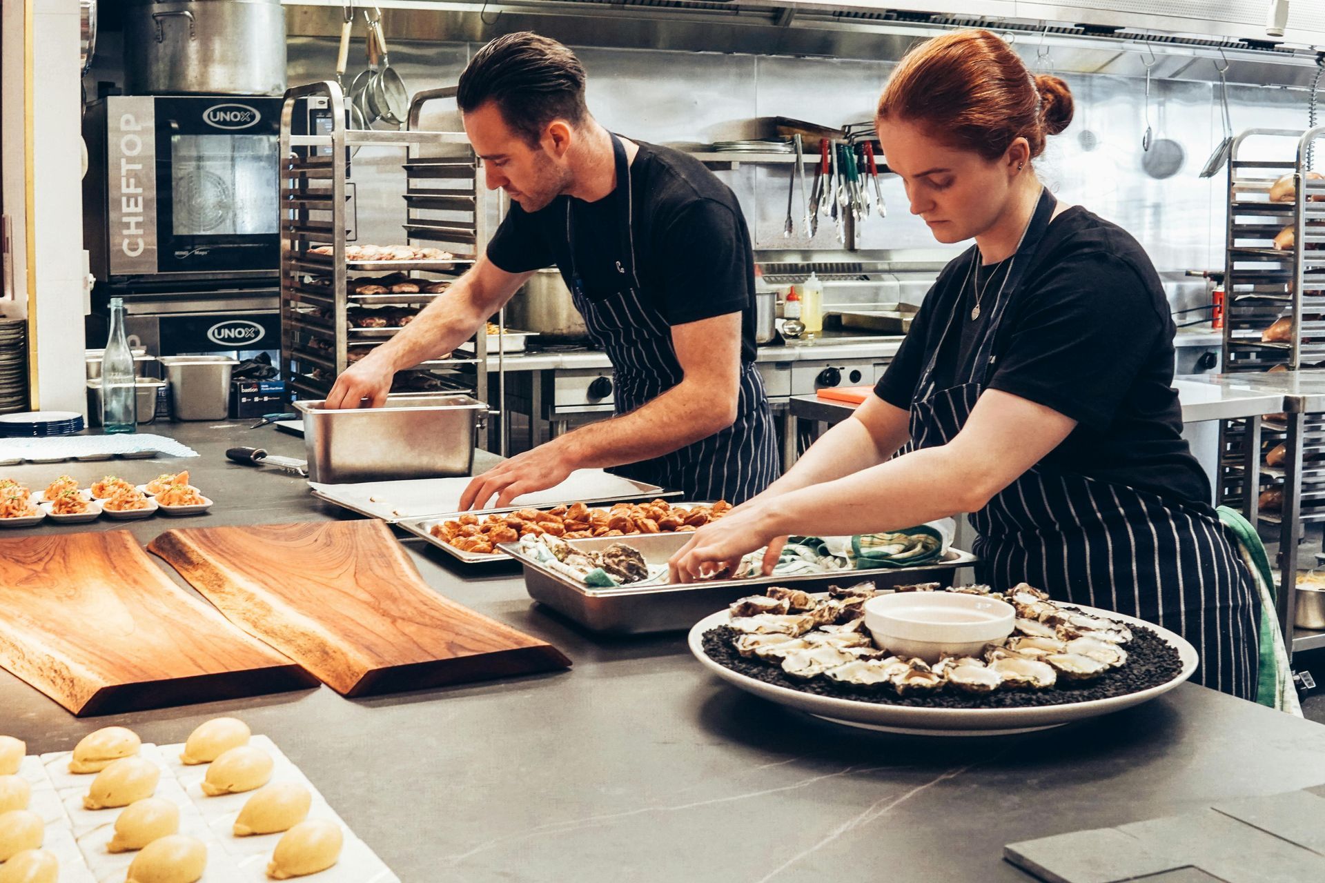 Two chefs preparing food in a commercial kitchen; one places items in a pan, the other adds food to a large plate.