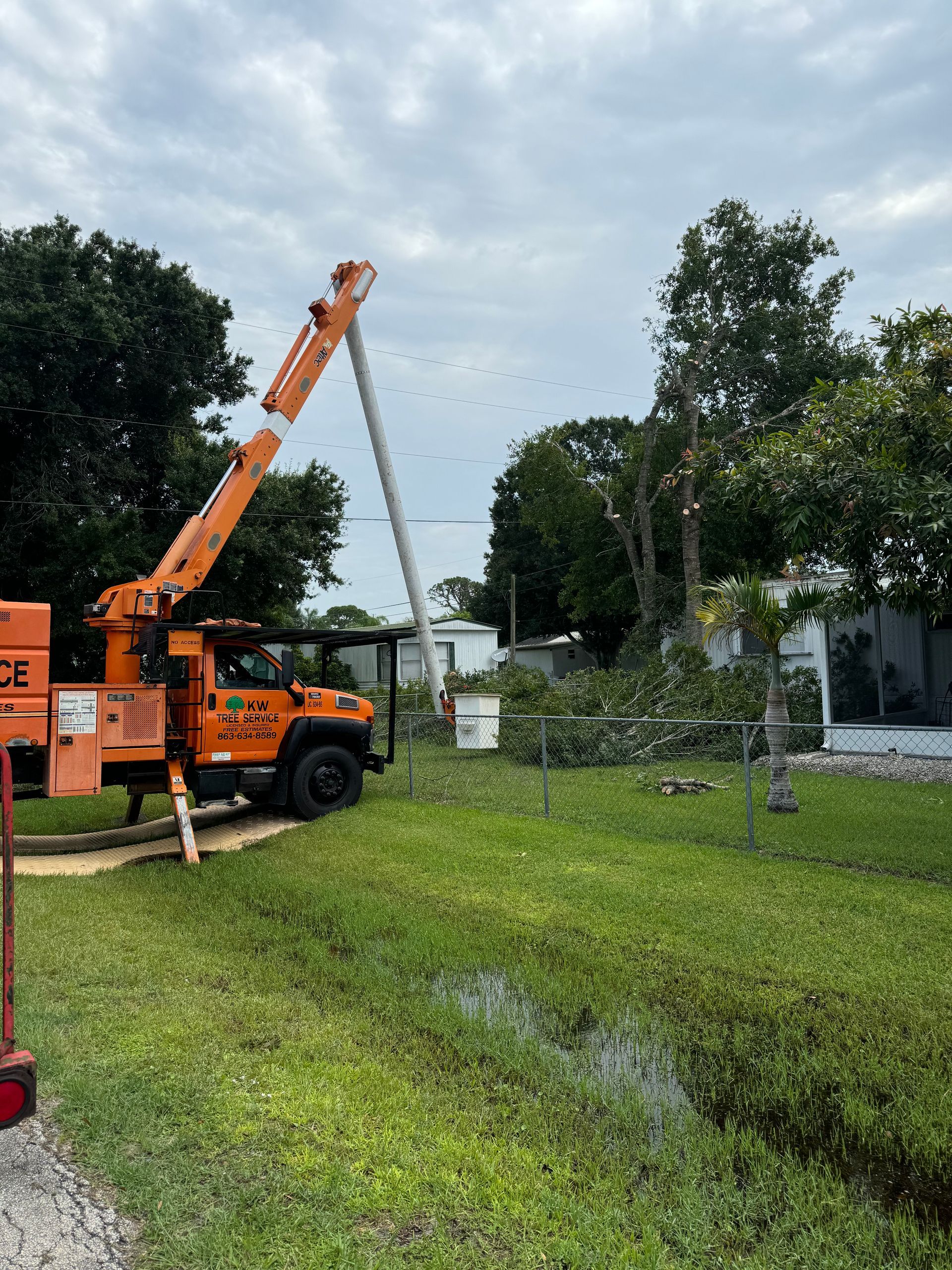 Orange crane truck lifting a long white pole in a grassy yard near trees and a house