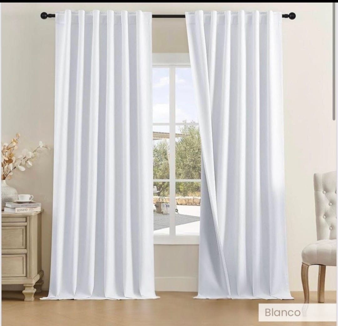 Cortinas blancas enmarcan una ventana, con una vista del exterior. Cerca hay una mesa y una silla.