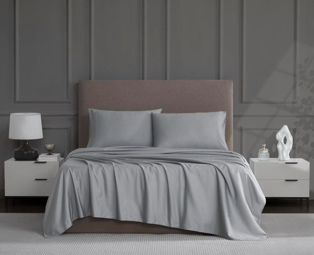 Sábanas grises en una cama con cabecero gris. Mesillas de noche a ambos lados con lámparas y adornos.