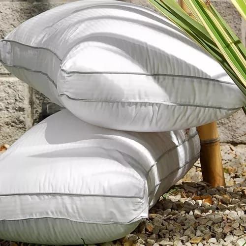 Dos almohadas blancas apiladas, contra una pared de piedra, con bambú y pequeñas rocas.