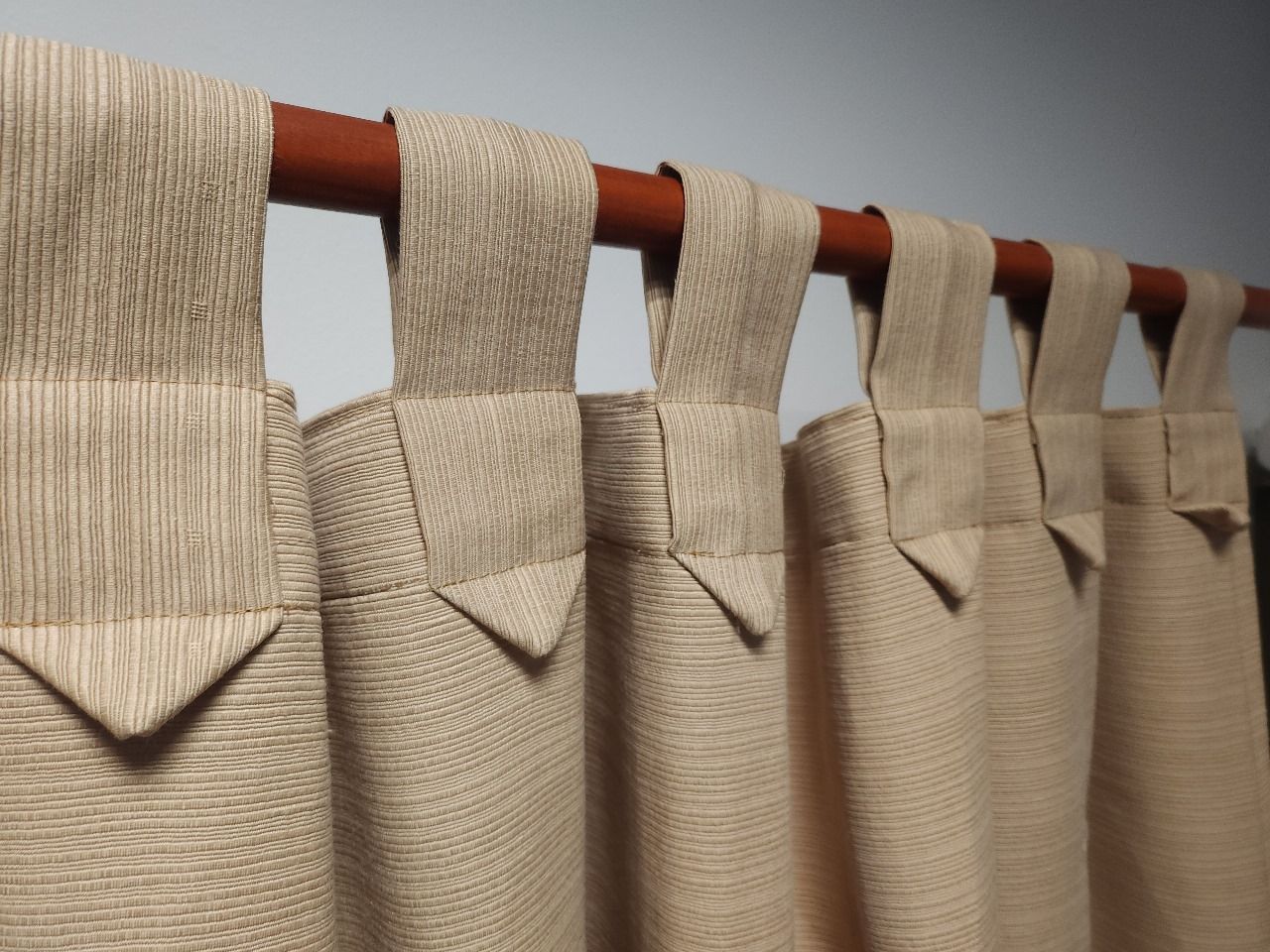 Cortinas de tela beige con trabillas que cuelgan de una barra de cortina marrón contra una pared blanca.