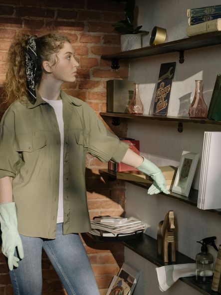 A woman wearing green gloves is cleaning a shelf in a room.