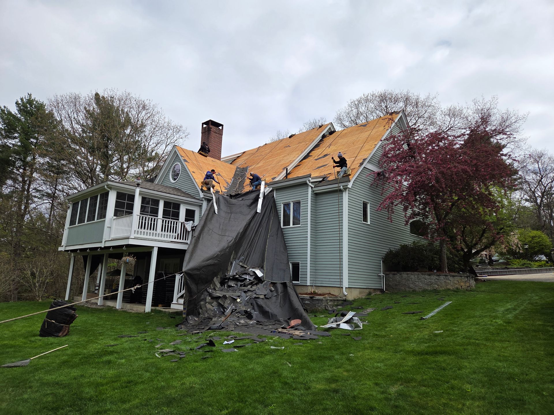 Damaged two-story house with torn roof and tarp-covered porch after storm, surrounded by trees.