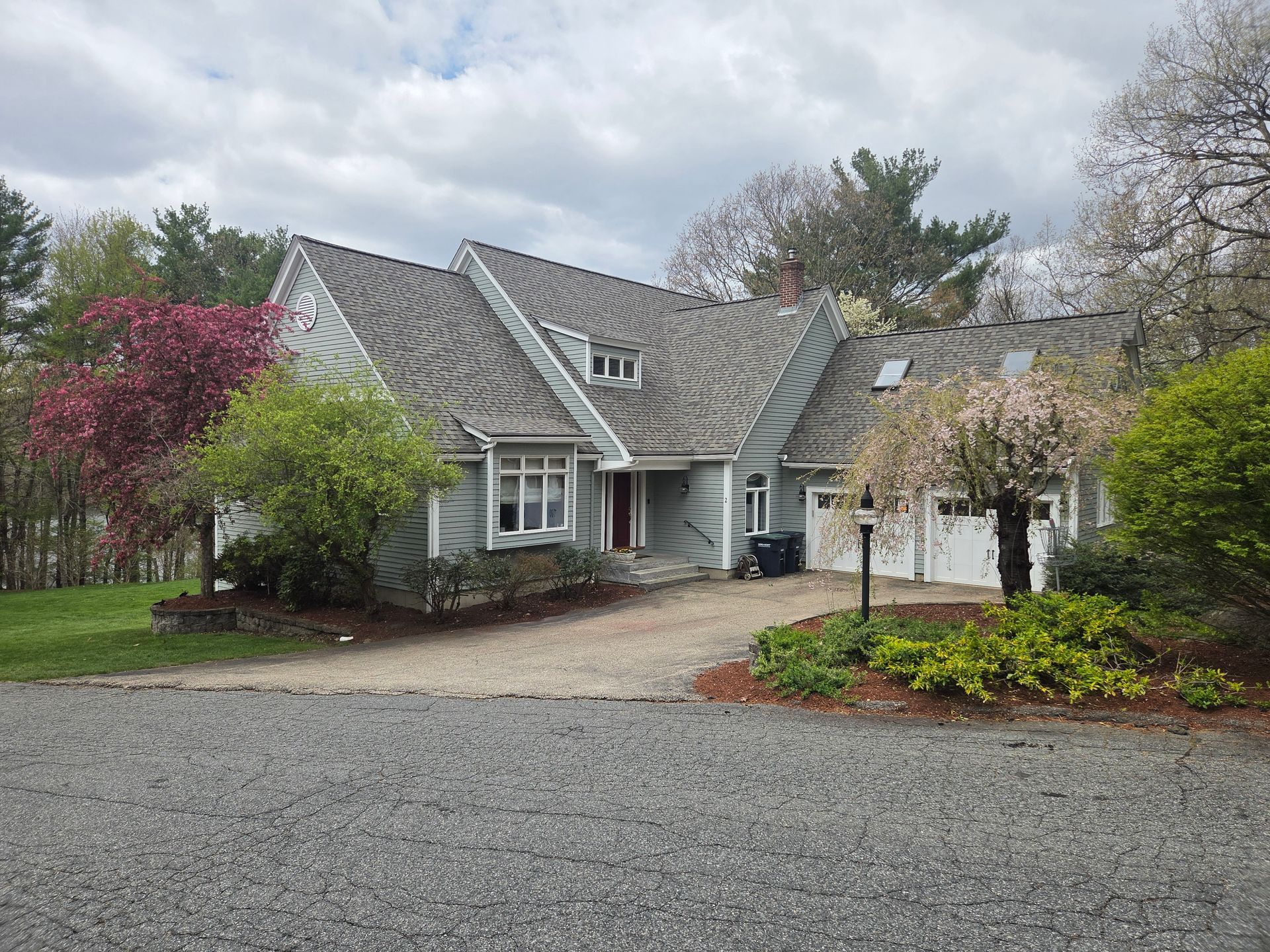 Gray house with gabled roof and driveway, surrounded by trees and landscaping under a cloudy sky