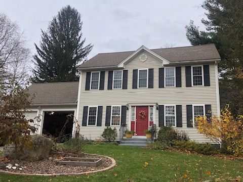 Two-story gray house with black shutters, red front door, and a garage, on a lawn with autumn foliage.