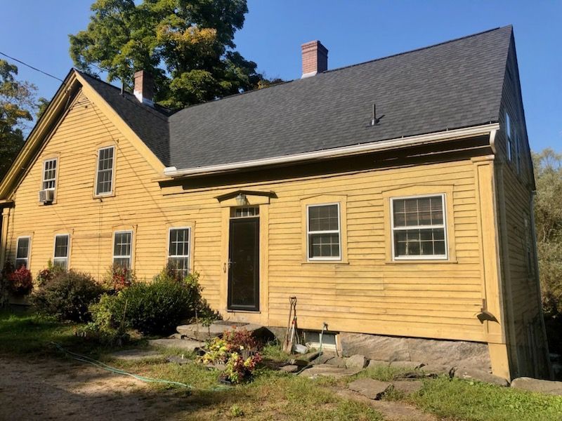 Yellow wooden house with black roof and stone foundation in a sunny yard