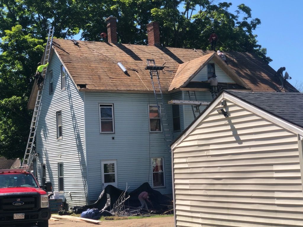 Two-story white house with roof damage, a red car parked beside it, and a small shed in front