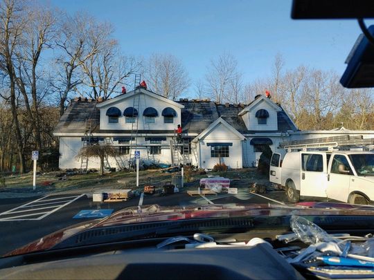 White house with dormer windows beside a parking lot, viewed from inside a car on a clear day.