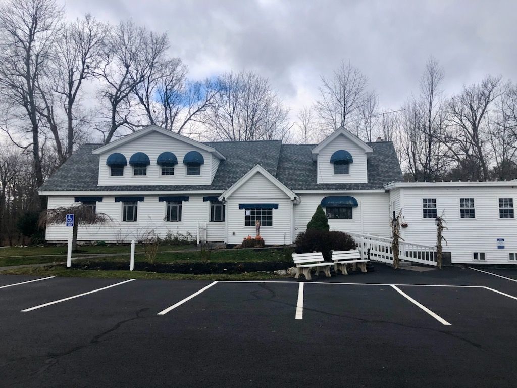 White building with green roof, dormers, and parked car in front of an empty lot under cloudy trees
