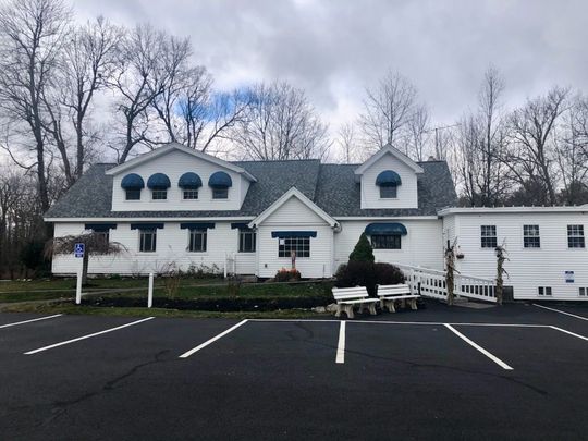 White building with gray roof and dormer windows, seen across an empty parking lot on an overcast day