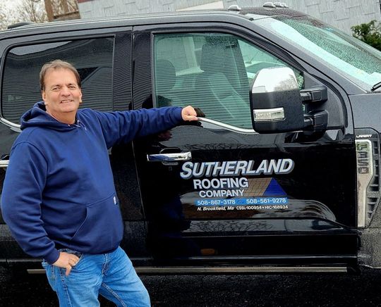 Man standing beside black Sutherland Roofing Company truck, wearing a blue hoodie and jeans.