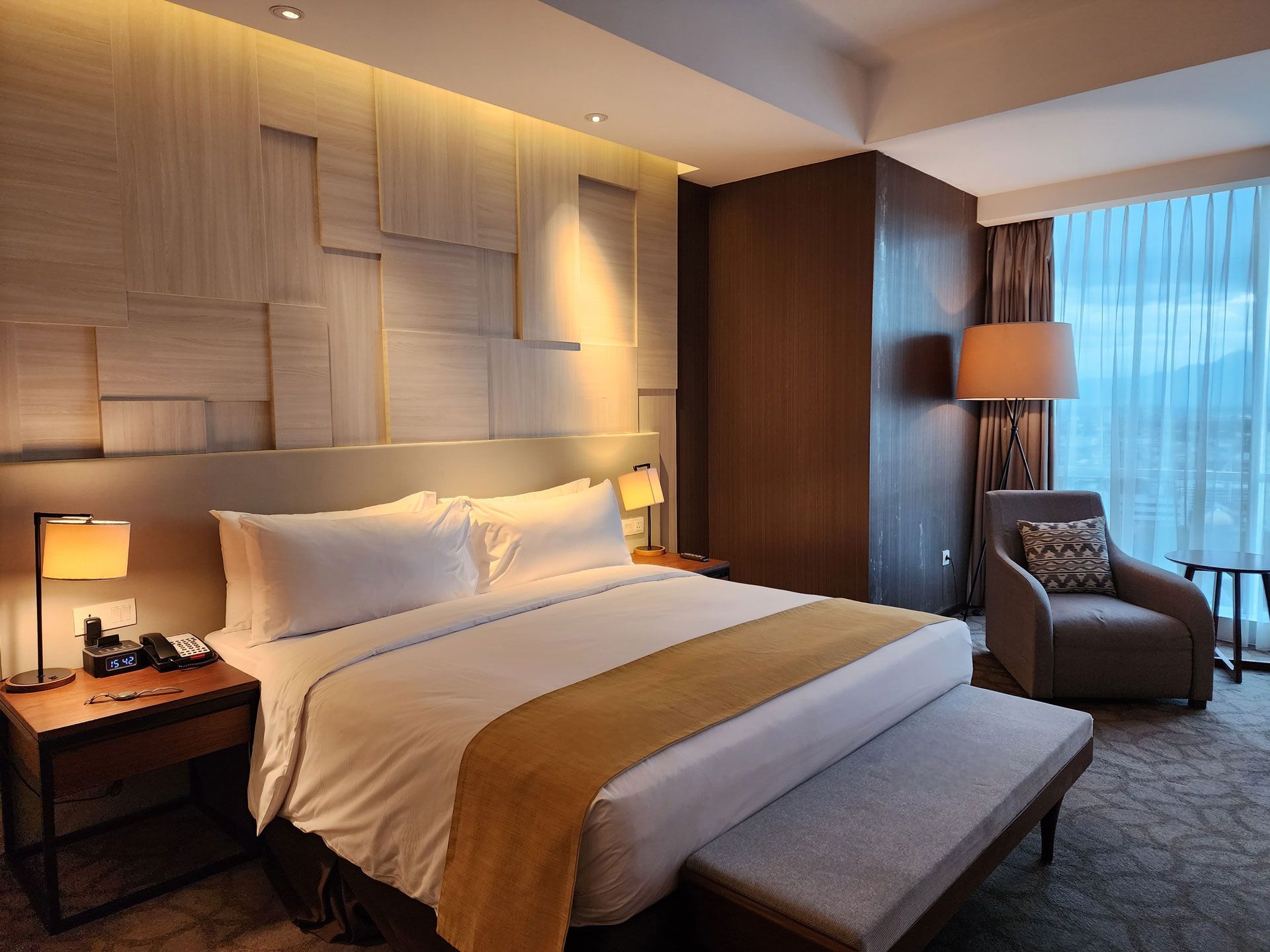 Hotel bedroom with king-sized bed, armchair, nightstands, window with curtains, and warm lighting.