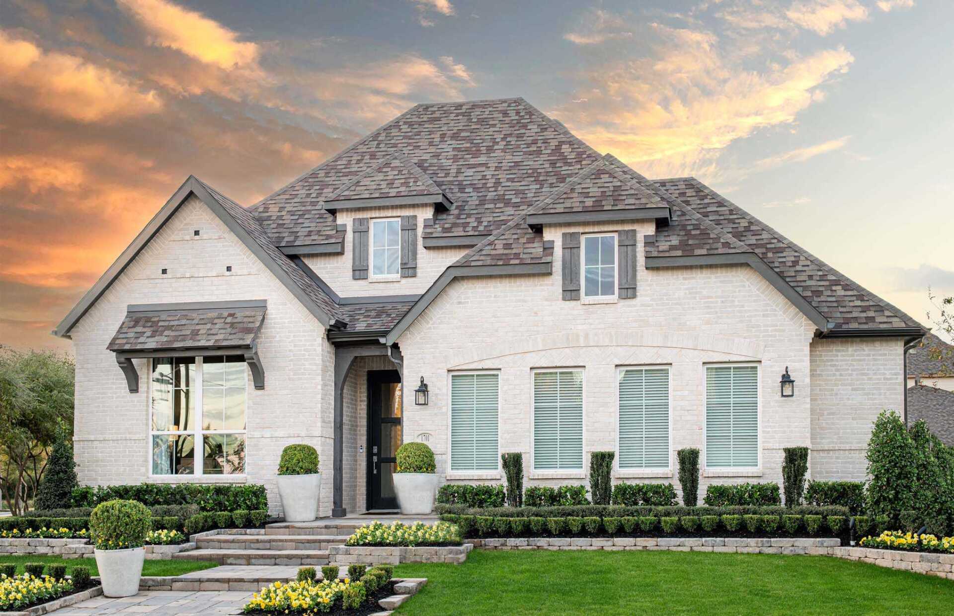 White brick home with gray roof, green lawn, and manicured landscaping under an orange sunset sky.