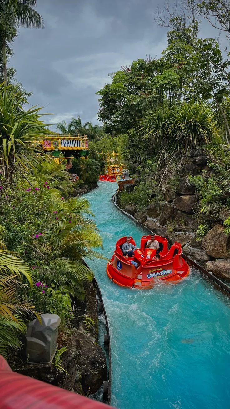 Botes vermelhos percorrem um toboágua turquesa em um parque de diversões. Uma vegetação exuberante cerca o canal.
