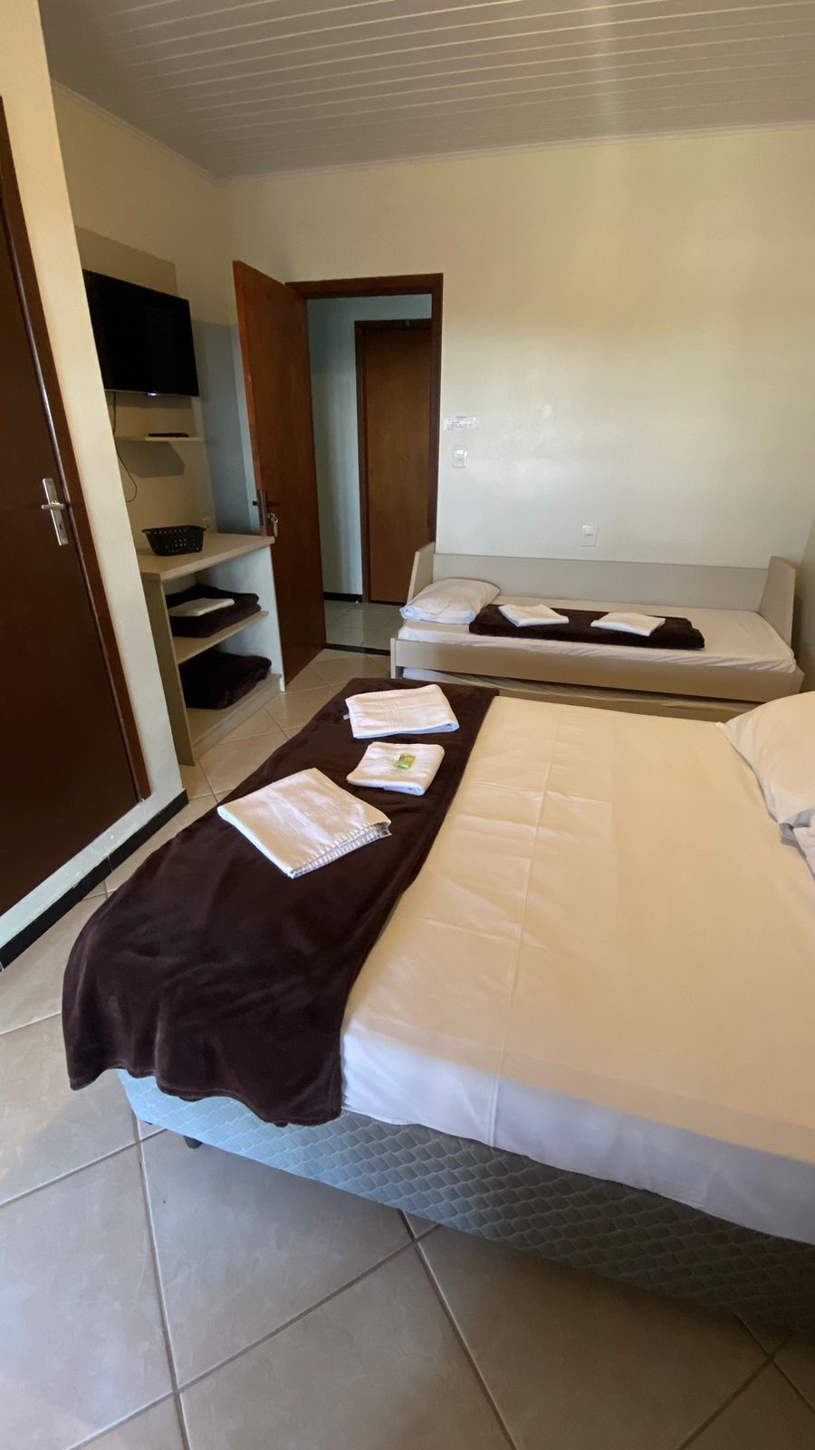 Quarto de hotel com cama, sofá, TV e porta aberta. Paredes bege, porta marrom, piso de azulejo.