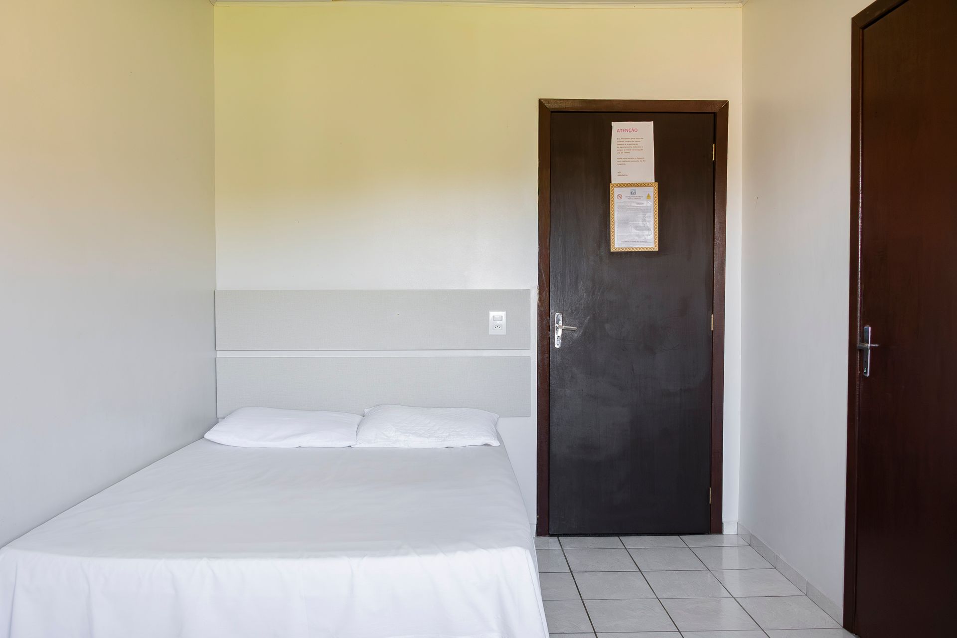 Quarto com cama de solteiro, roupa de cama branca e porta marrom escura. Paredes brancas e piso de azulejo.