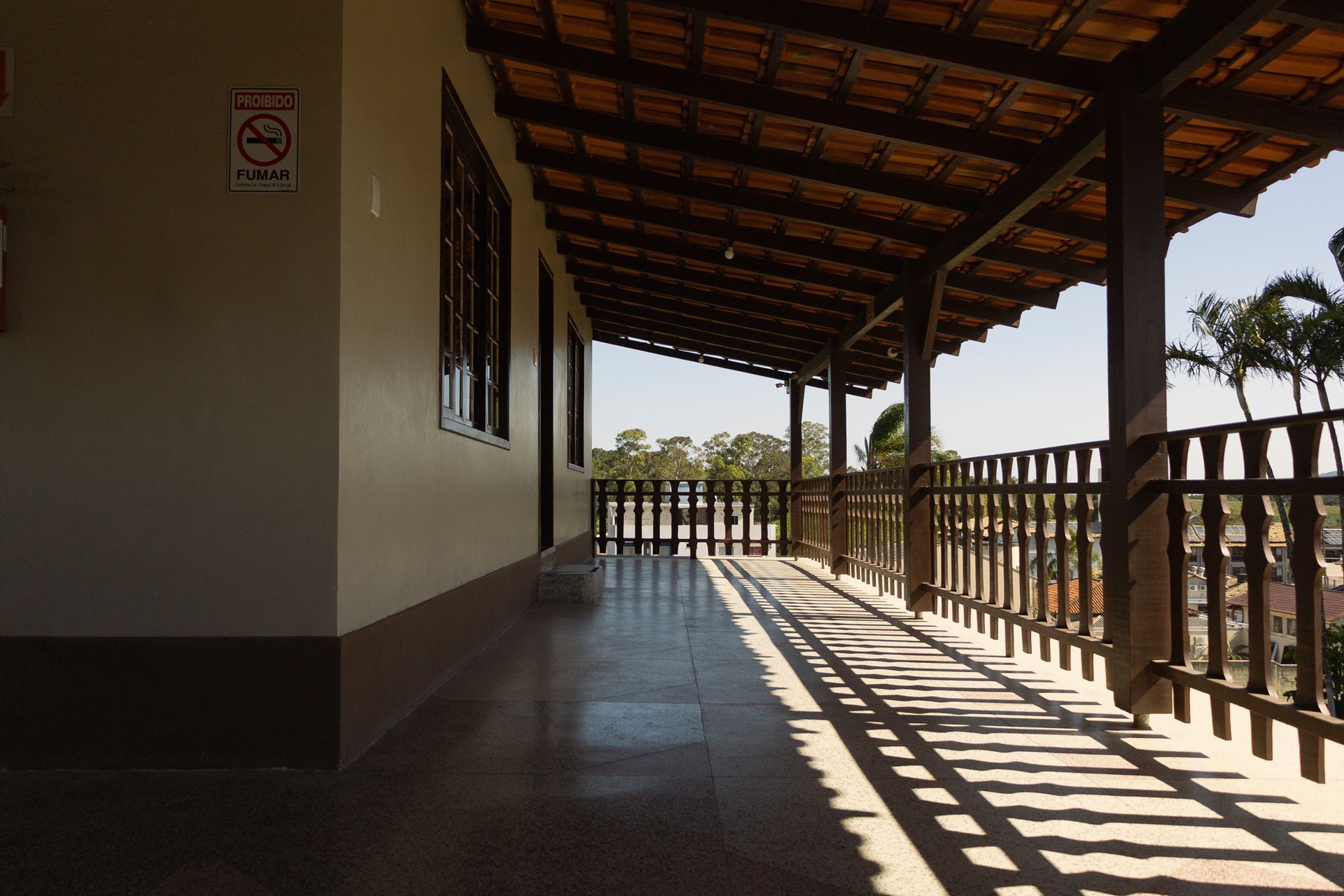 Passarela externa coberta com corrimão de madeira e telhado de telhas, projetando sombras no chão.