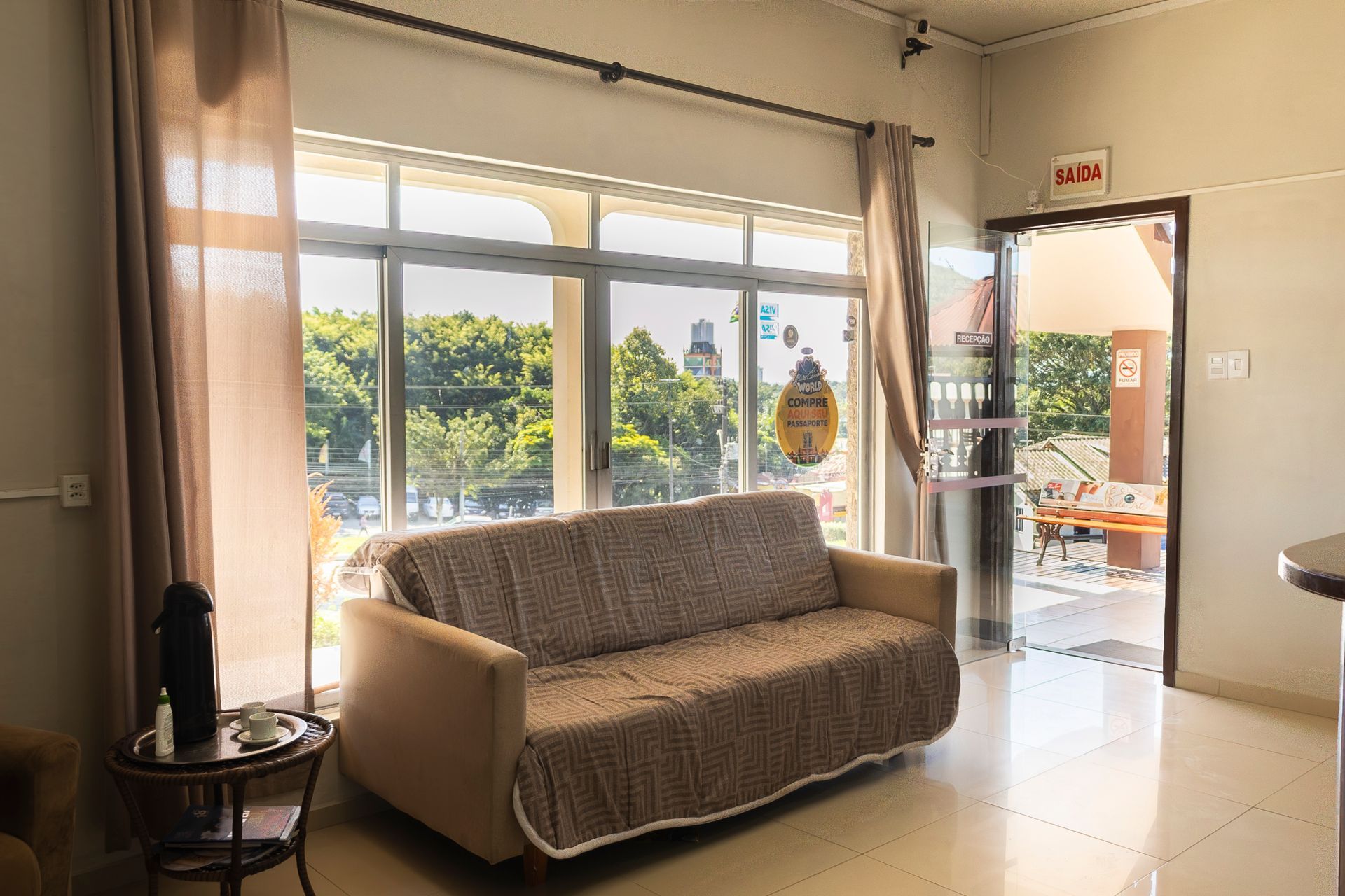 Sala de estar com sofá coberto por uma manta estampada, janela grande e porta aberta para um pátio.