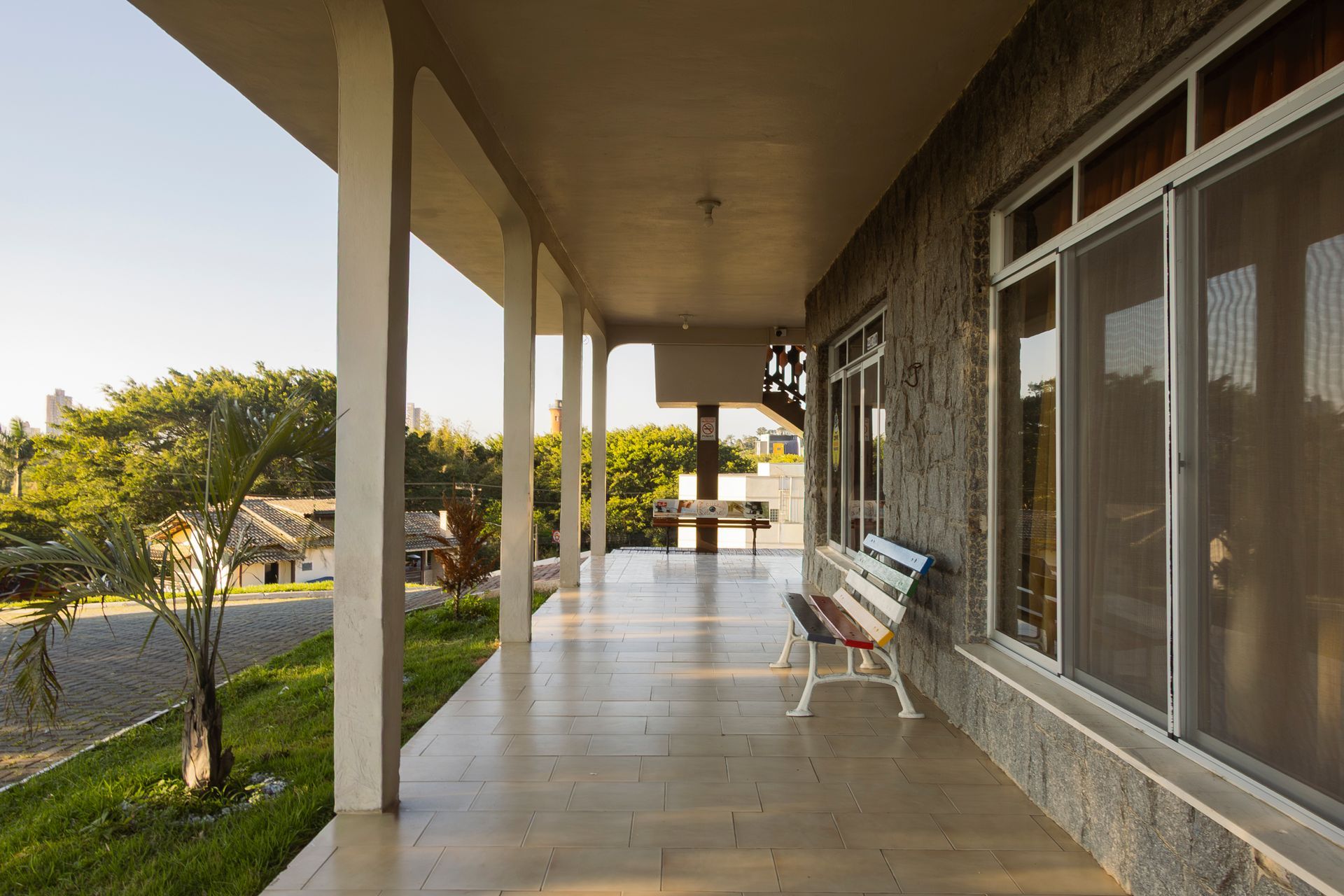 Uma longa varanda coberta com colunas, um banco e janelas em um edifício com vista para as árvores.