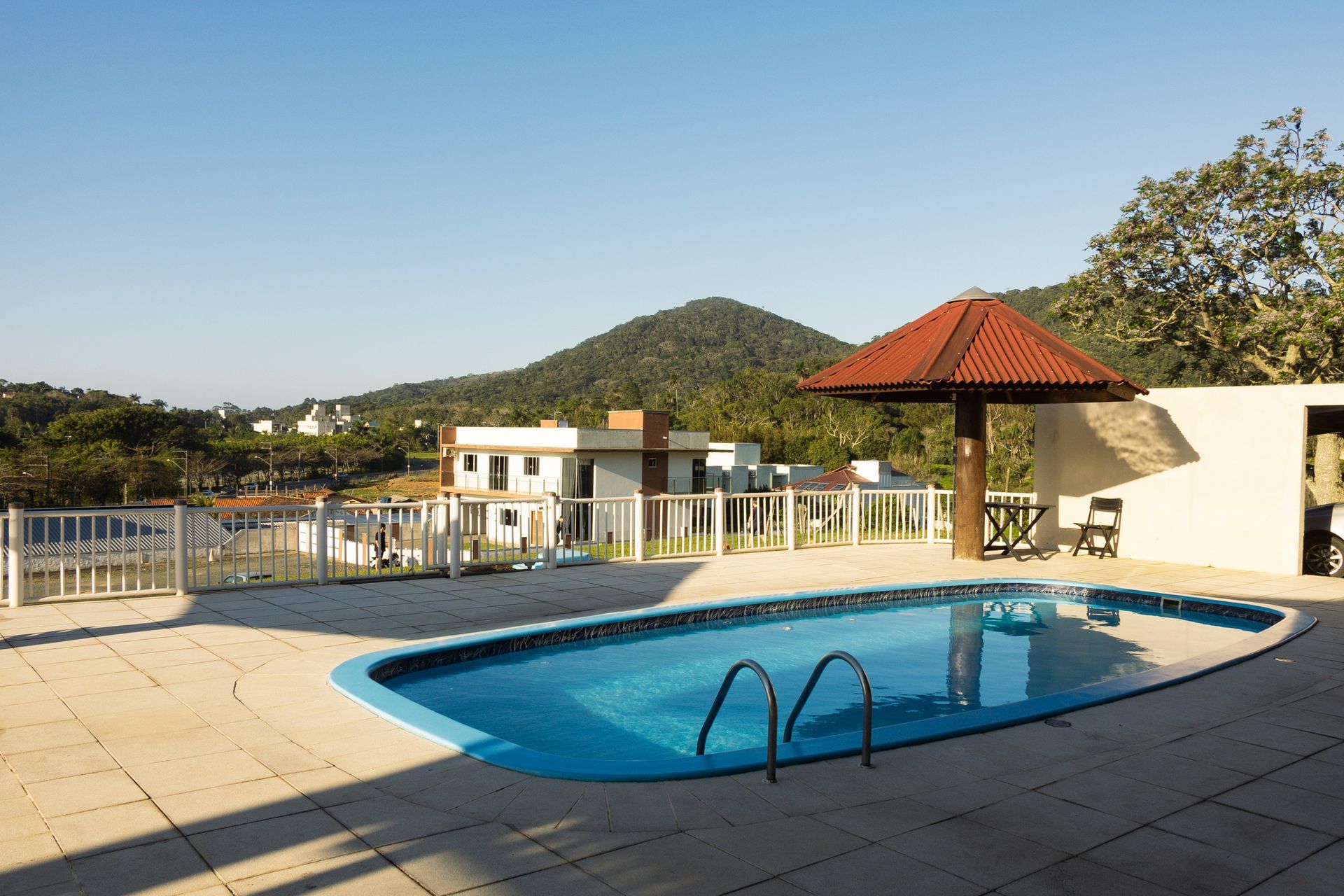 Piscina com vista para a montanha, sob uma estrutura com telhado marrom, em um dia ensolarado.